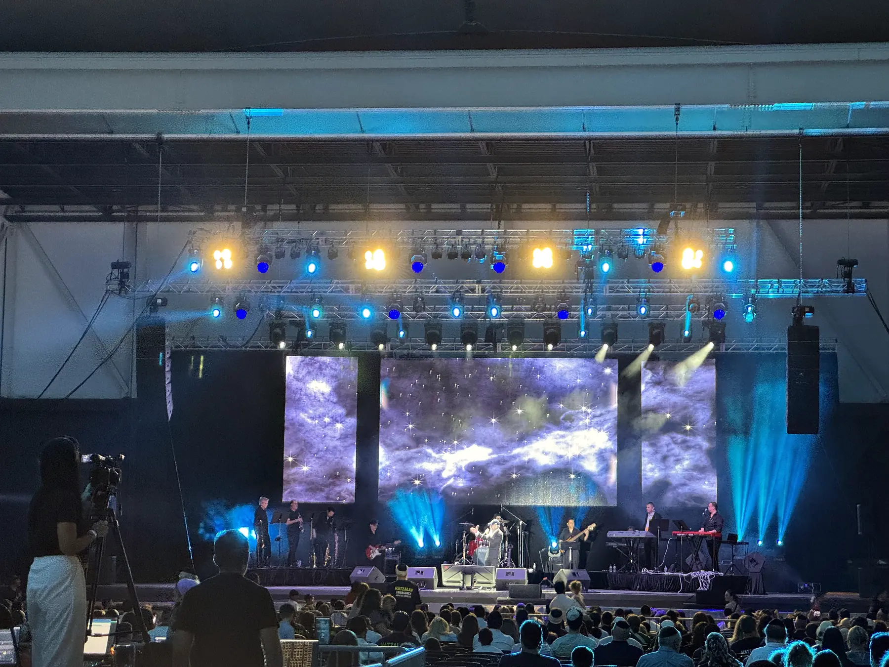 A concert under a high roof, three tall video panels behind the band carrying one image across them, two rows of lighting fixtures hung on truss above, and an audience filling the floor in silhouette.