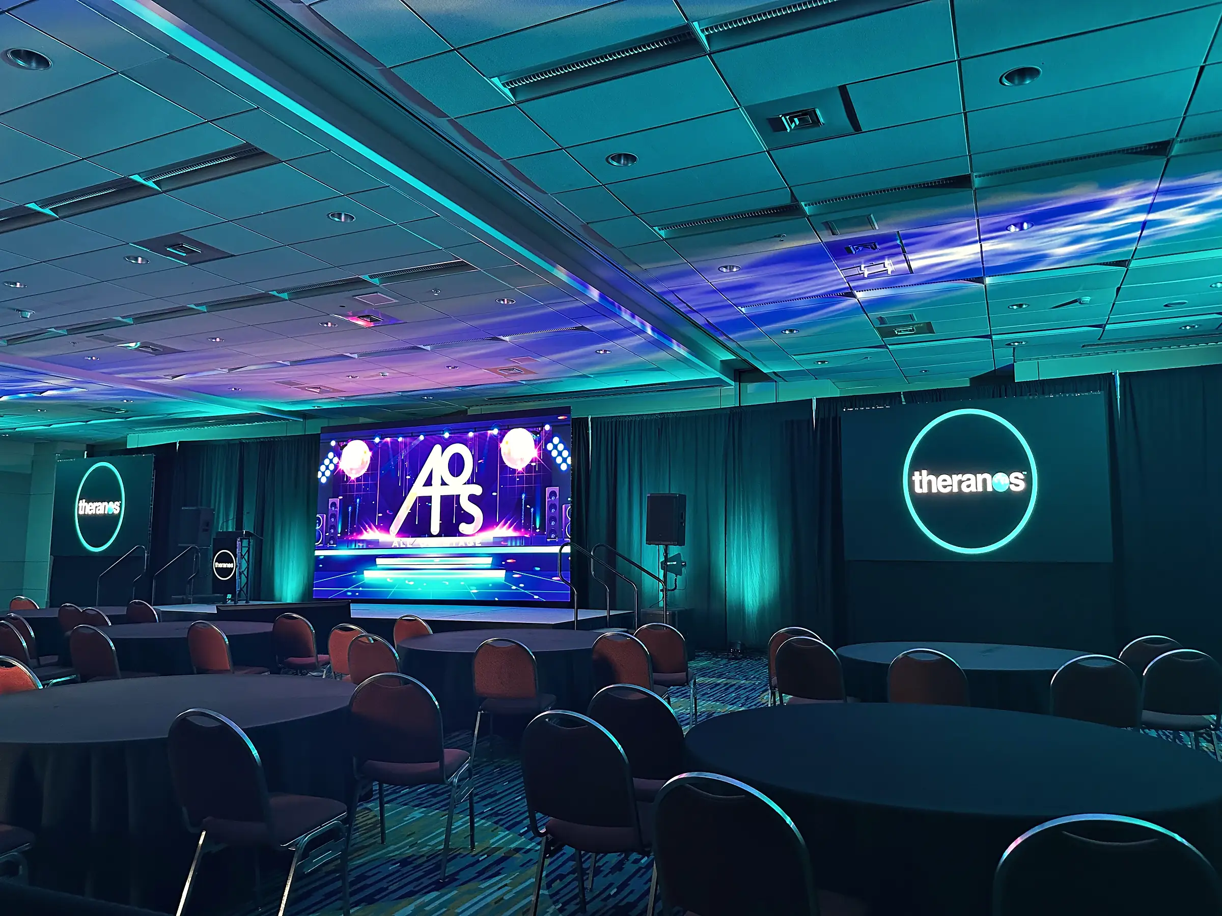 A ballroom washed in cyan and blue with rounds set in front of a low stage, a video wall upstage carrying a graphic, a second large screen filling the side wall, and lit rings hung on the drape between them.