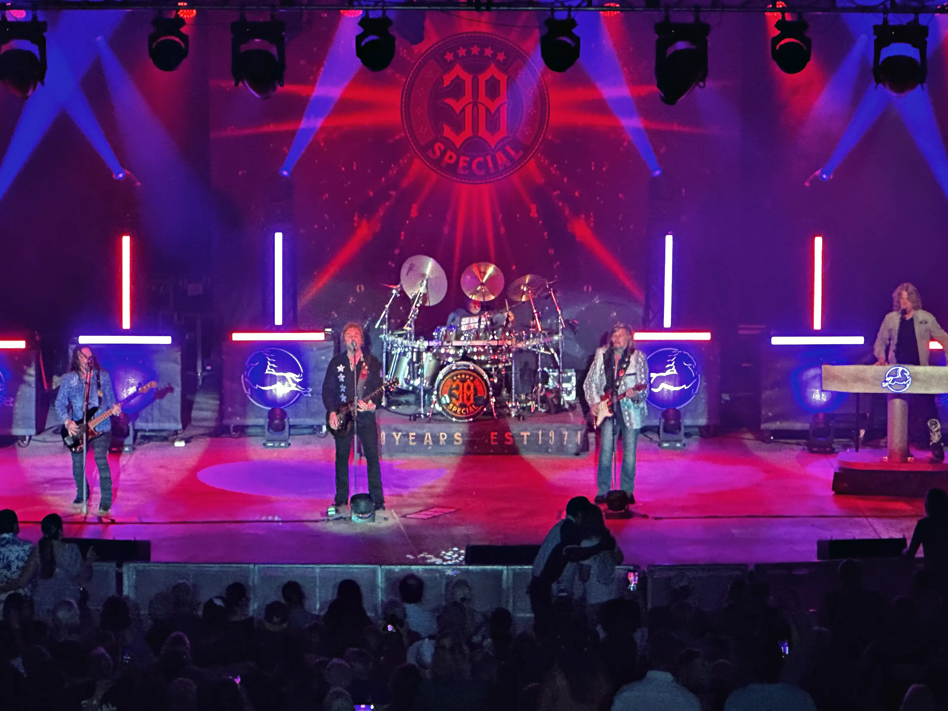 A band on a stage lit red and blue, a drum kit on a riser at the centre with guitar and keyboard positions either side of it, vertical light bars and a scenic wall behind them, and the heads of an audience across the bottom of the frame.
