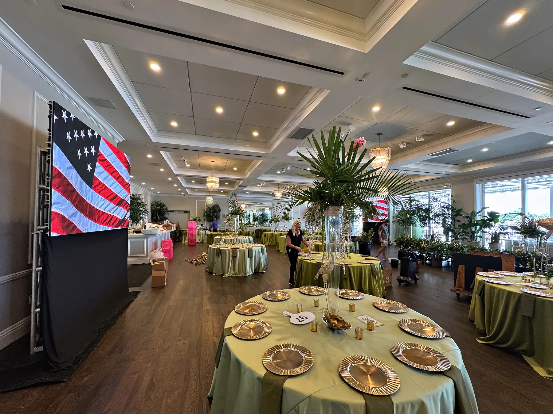 Banquet rounds set with pale green linens, gold chargers and tall foliage centerpieces in a paneled room, a large flag displayed on a raised panel at the left and daylight coming through windows at the back.