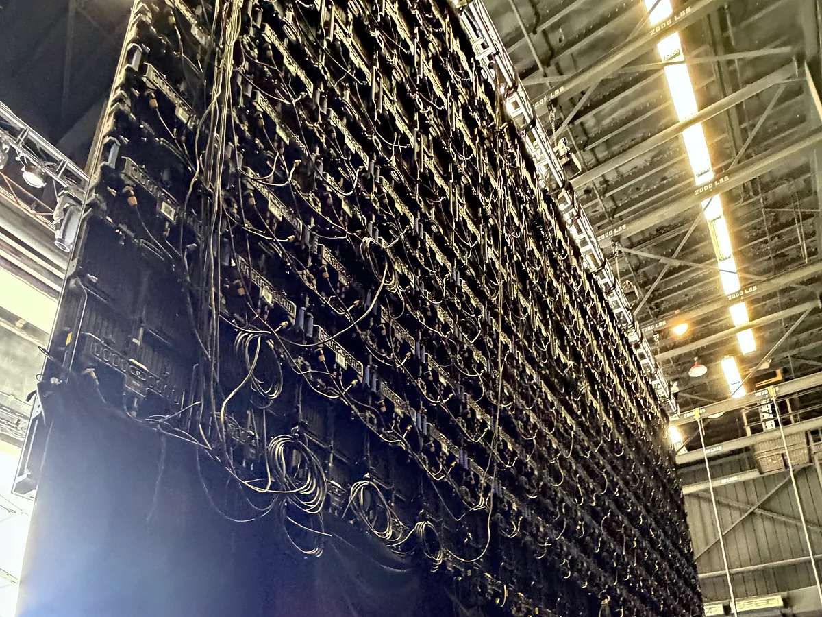 A dense row of chain hoists and their chain bags hanging from truss high in a room, black drape falling away below them and the lit ceiling running back behind.