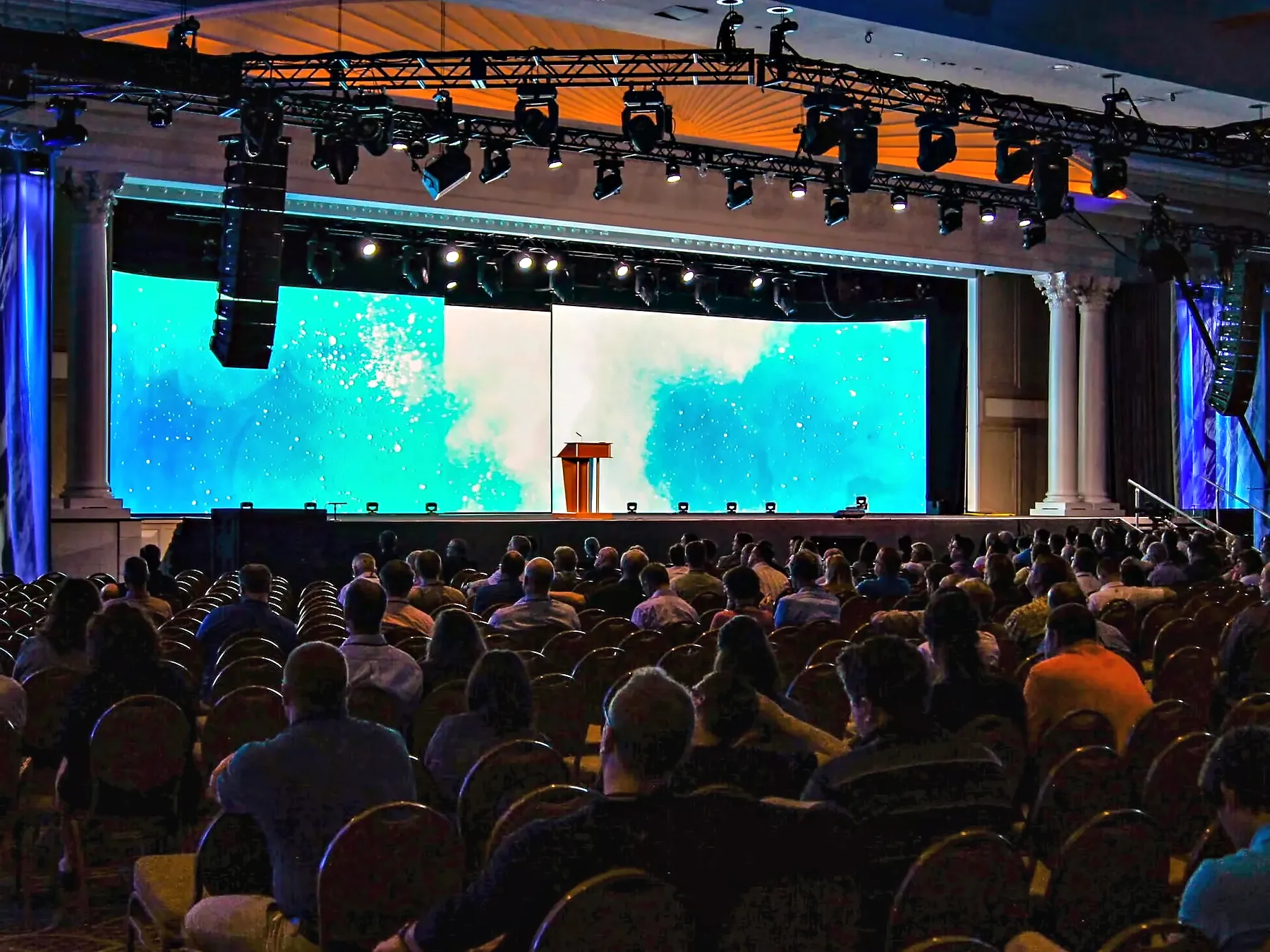 A full house seen from the back of the room, every seat taken, facing a wide video wall carrying an abstract image with a lectern lit at its center, loudspeakers flown at both sides and a lighting truss across the top of the proscenium.