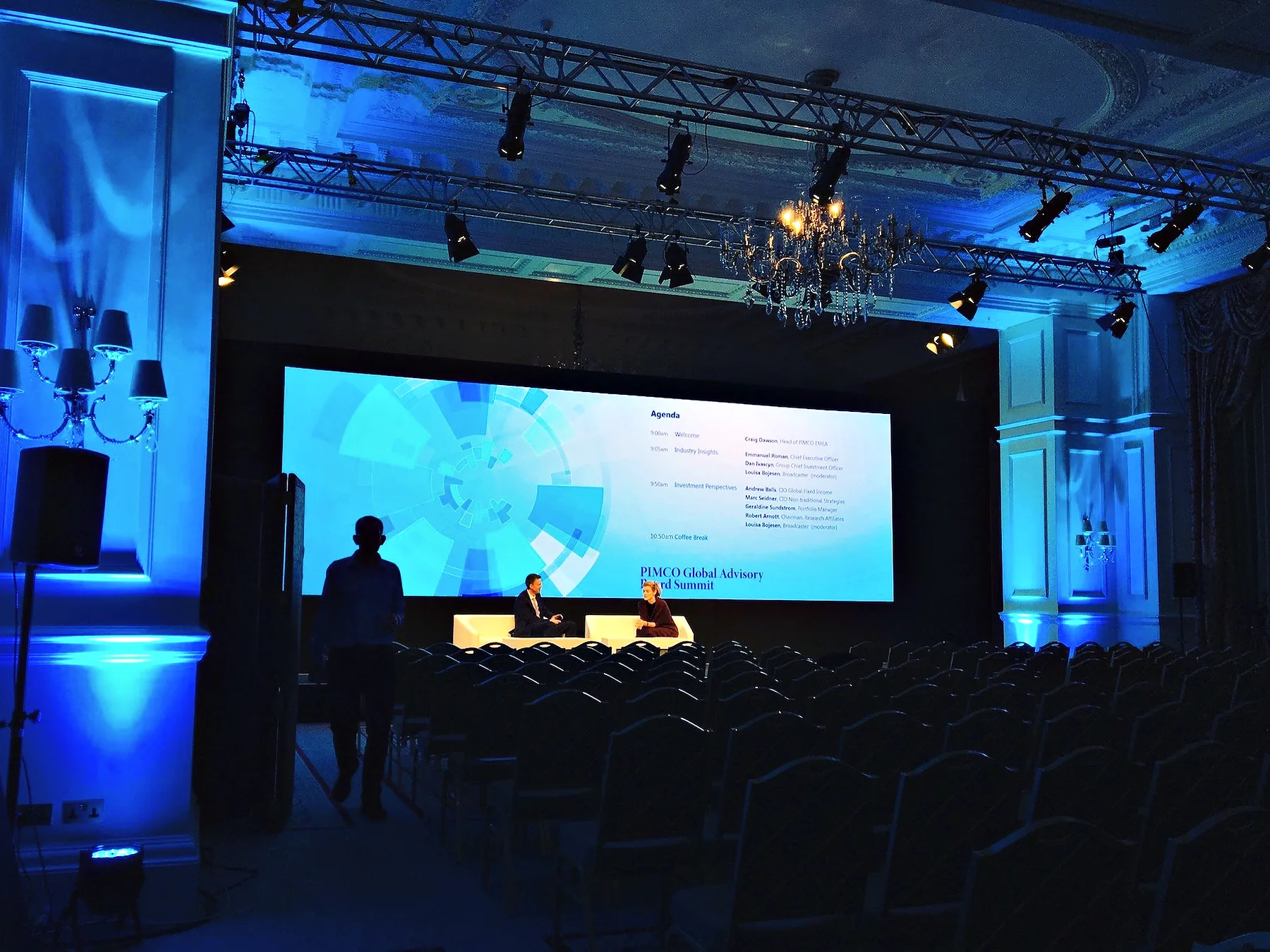 A darkened ballroom washed in blue, two people seated in armchairs on a low stage in front of a wide screen carrying a slide, lighting truss overhead and rows of empty chairs running back from the stage.