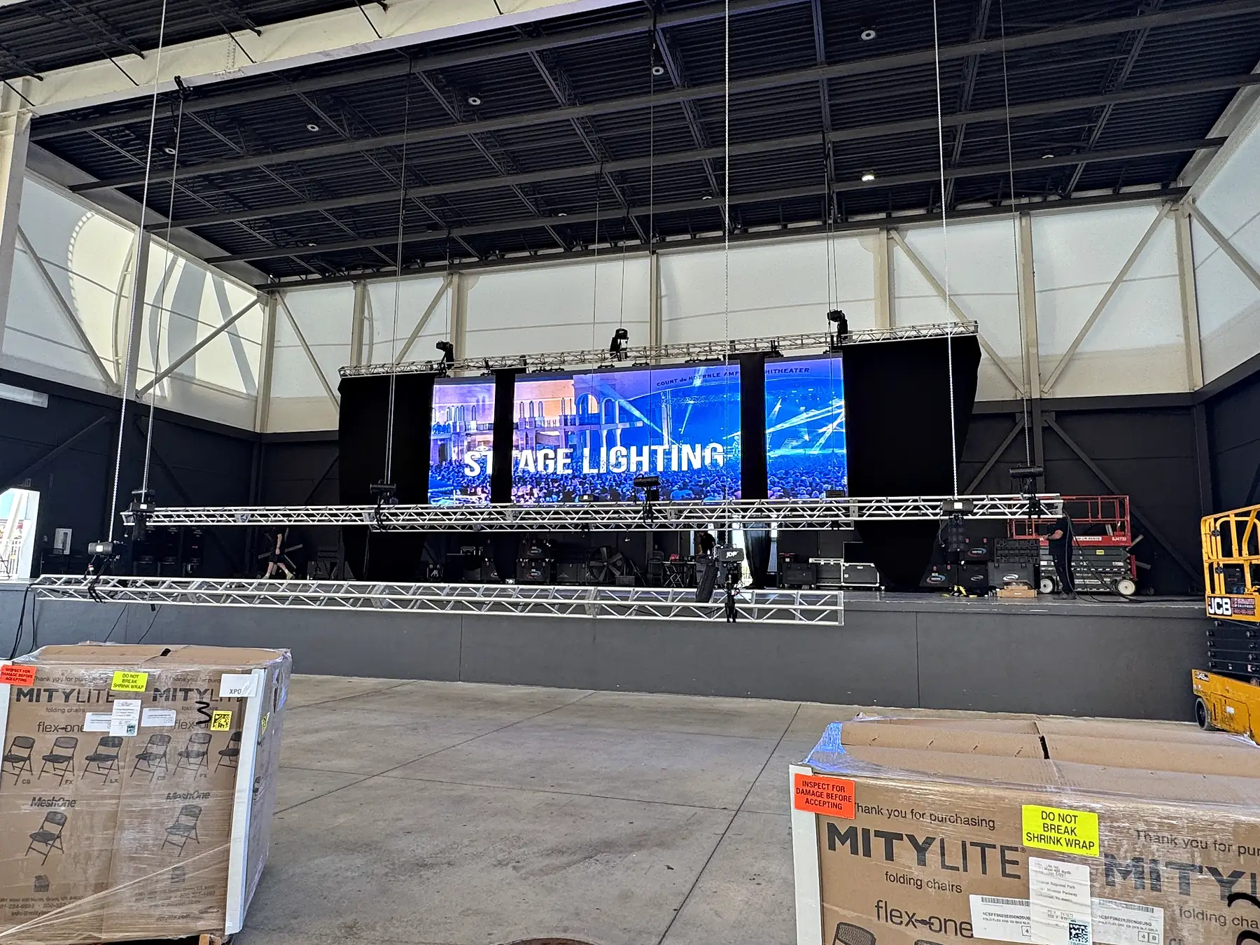 A stage built against the wall of an empty hall on ground-support truss, a video wall across the back of it on a test image, lighting fixtures hung on the front truss and pallets of stacked chairs still on the floor.