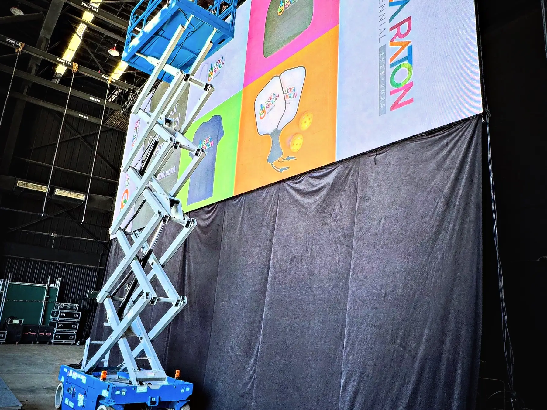 A tall LED wall carrying a full-bleed graphic above a draped base in a hangar-like room, a scissor lift raised alongside it to the top edge, and a distribution rack standing on the floor at the right.
