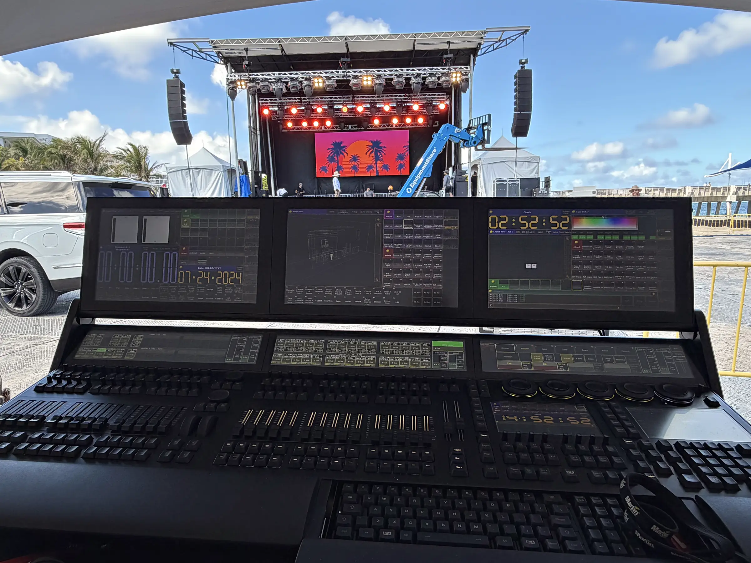 A lighting control surface in the foreground with its faders, wheels and displays lit, and beyond it an outdoor stage under a mobile roof with loudspeakers flown at both sides against a clear blue sky.