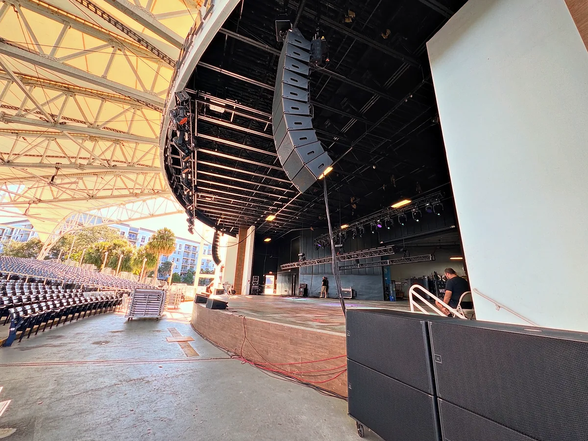 A line array hung close to the camera under the steel roof of a covered venue, the cabinets curving away and downward toward the stage, with rows of empty seats and daylight beyond them.