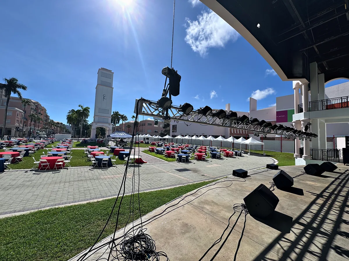 Loudspeakers standing on a brick plaza with coils of cable run between them in bright sun, a curved canopy overhead, and rows of tables under white tents set out across the lawn beyond.