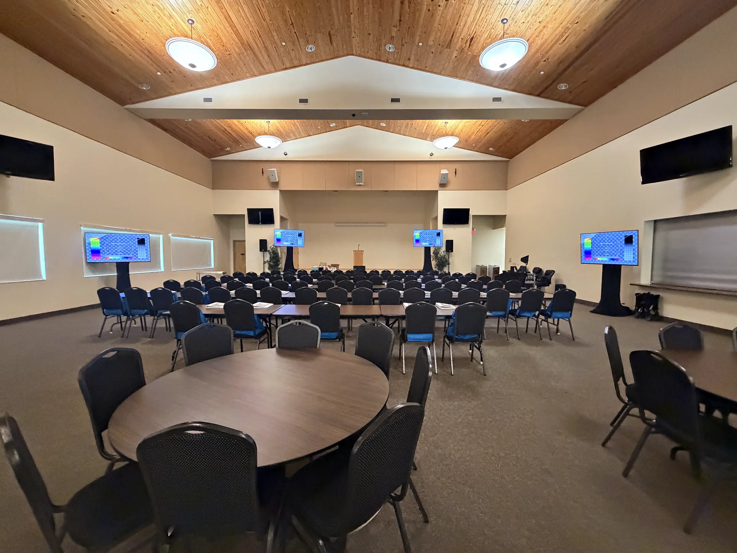 A meeting room under a pitched wooden ceiling set with round tables and stacking chairs, a lectern at the front and small wall-mounted screens carrying the same holding slide at each side.