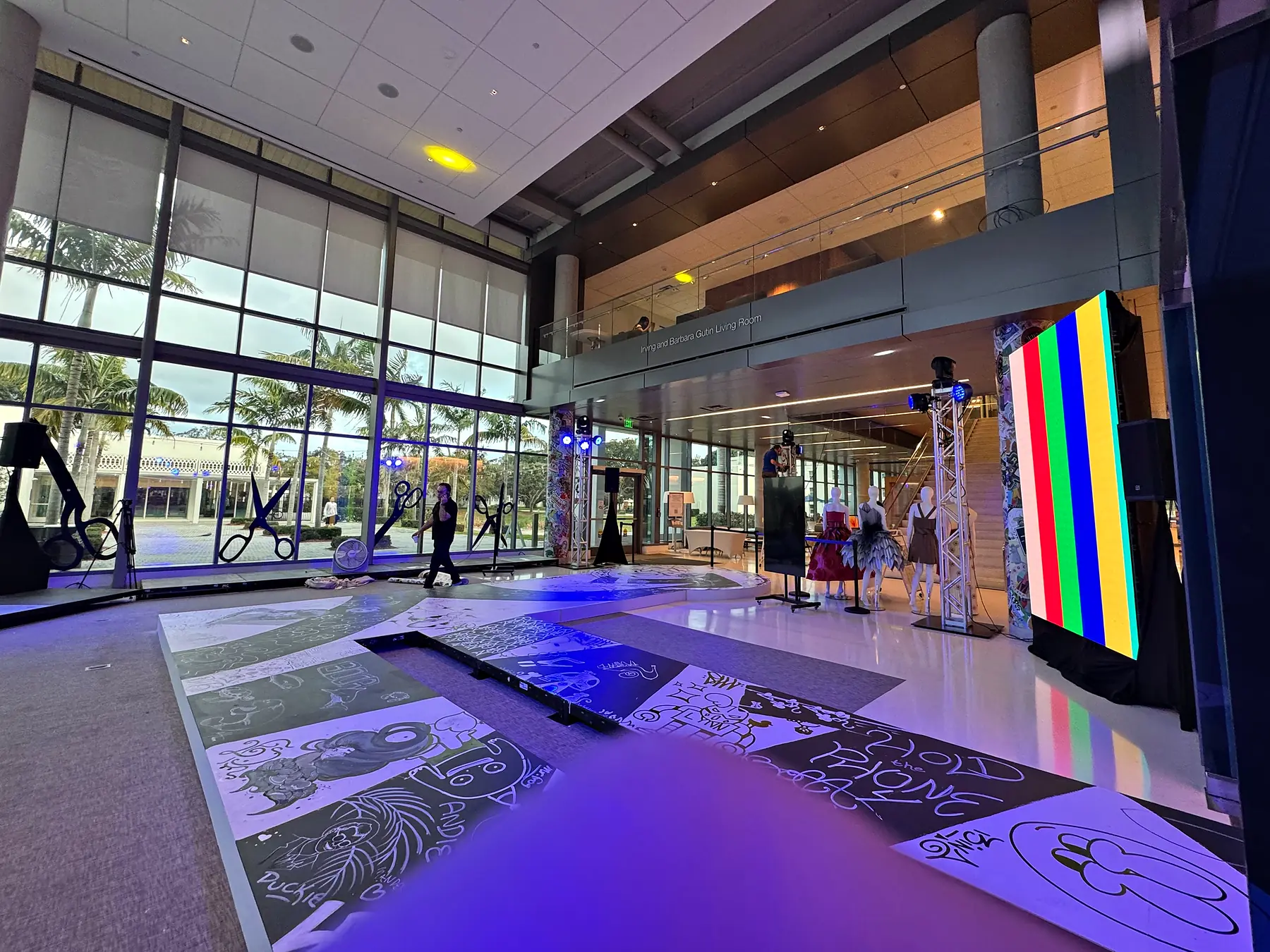 A runway laid down a glass-walled atrium with a printed graphic floor, tall portrait video panels carrying color bars at the far end, purple uplight across the space and daylight through the windows behind.