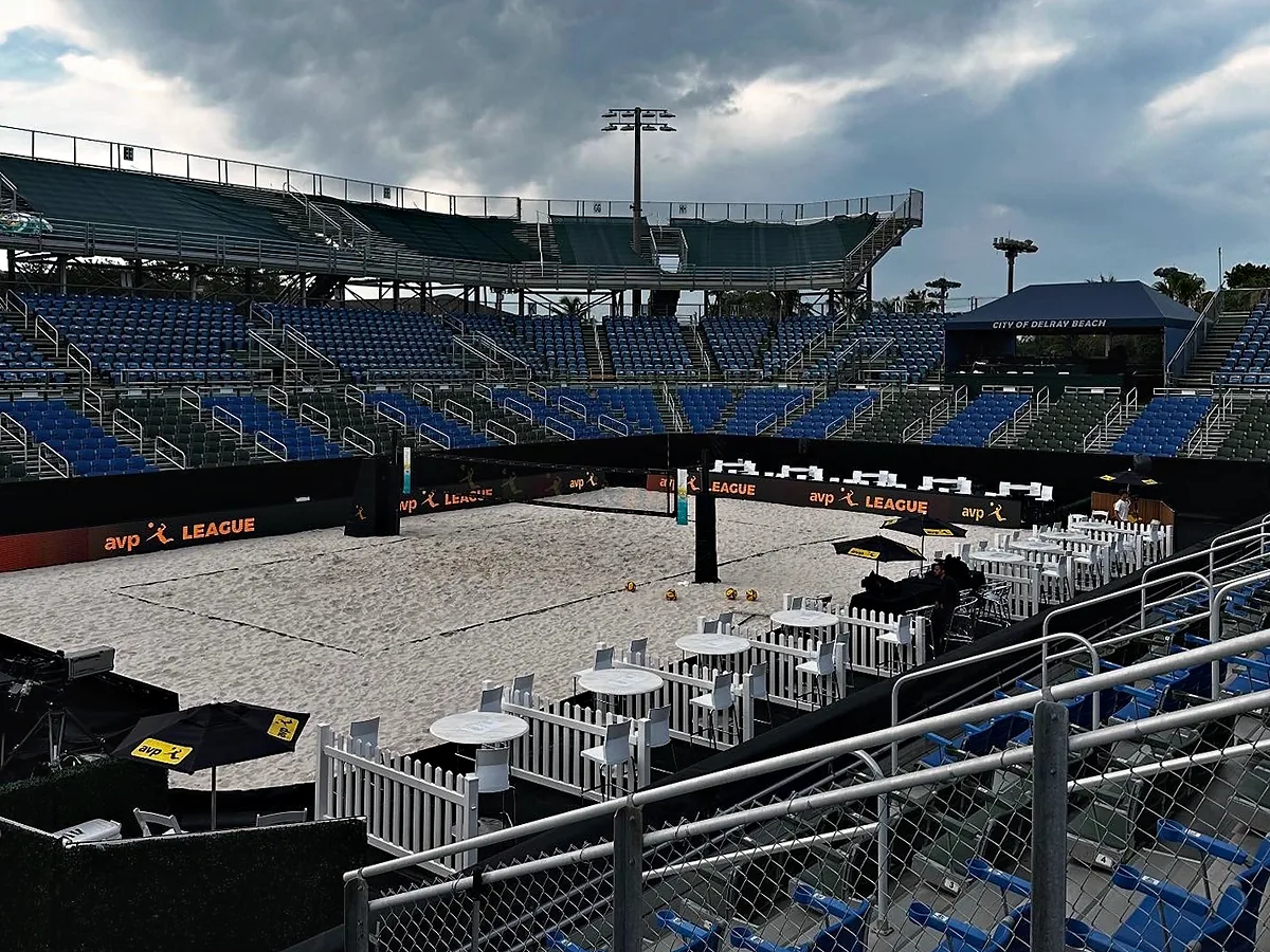A sand court laid out on the floor of a stadium bowl under a heavy overcast sky, temporary seating and railings ringing it, and the permanent stands rising empty behind.