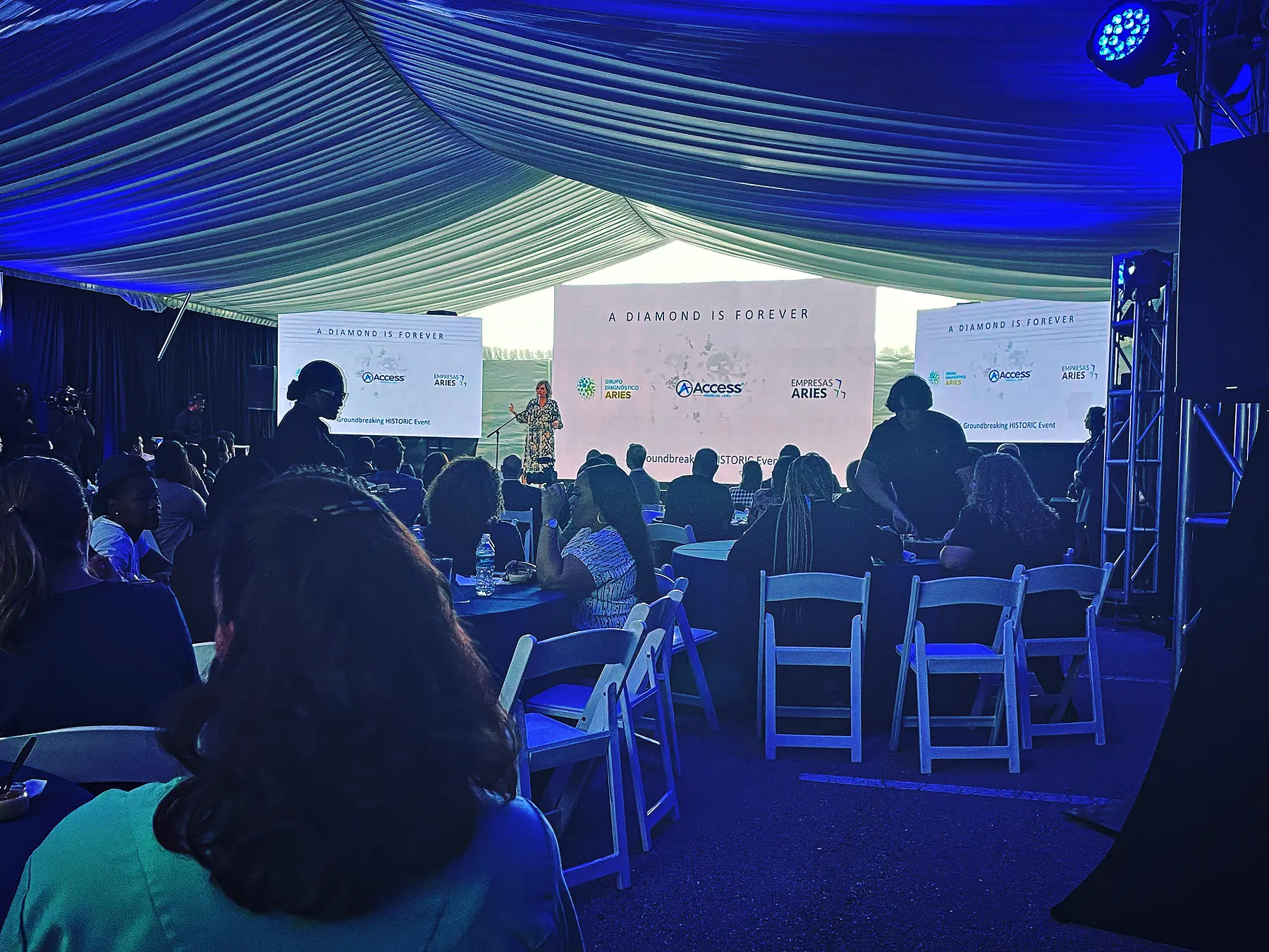 A tent with fabric draped in swags across its ceiling and lit blue, three screens across the far end of it carrying the same image, a speaker at a lectern between them and guests seated at round tables in front.