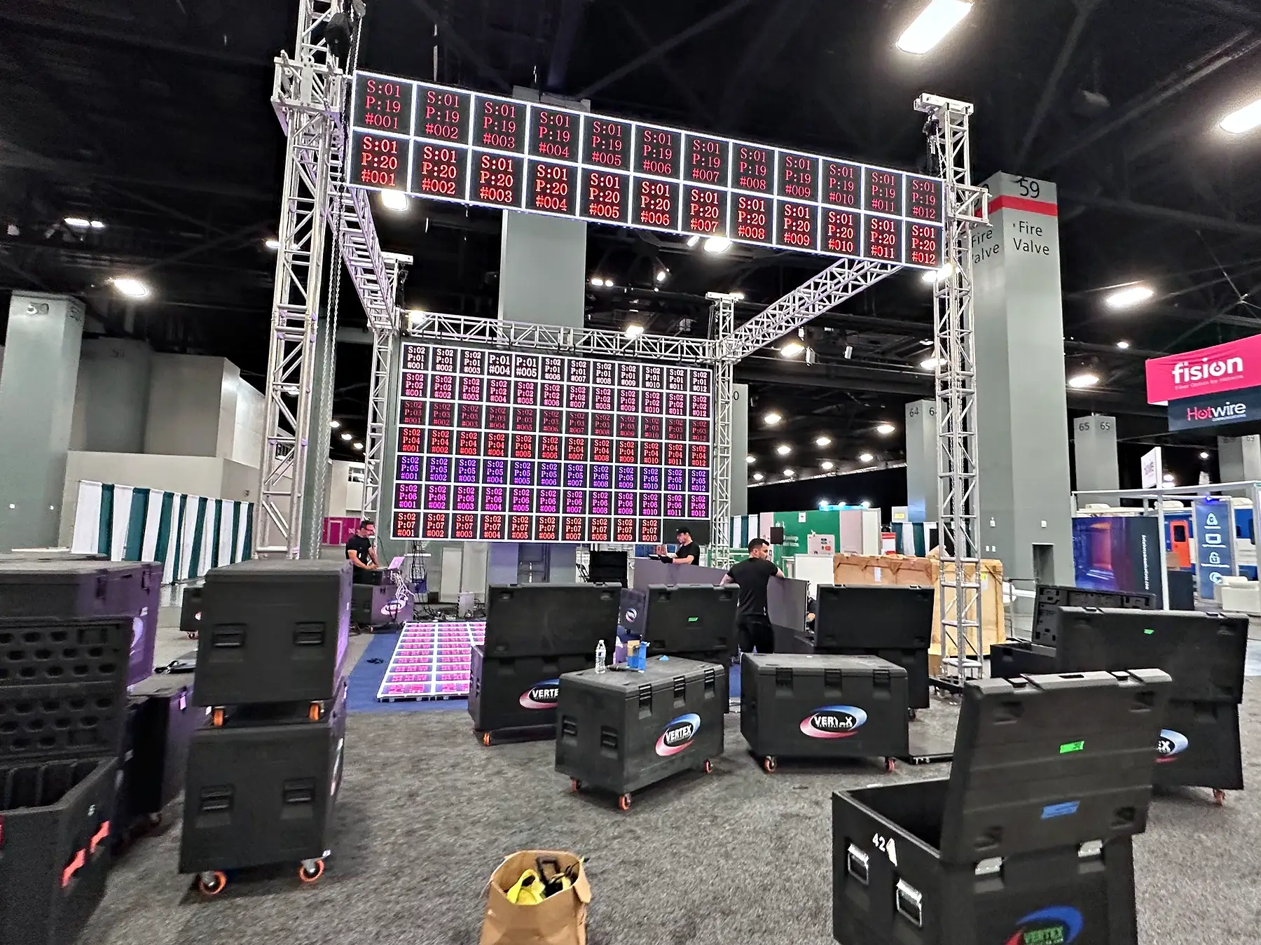 A trade show booth mid-build on a convention floor: a square of ground-supported truss carrying LED panels on a red and white test field, loudspeakers and road cases stacked on the concrete around it and the hall running away behind.
