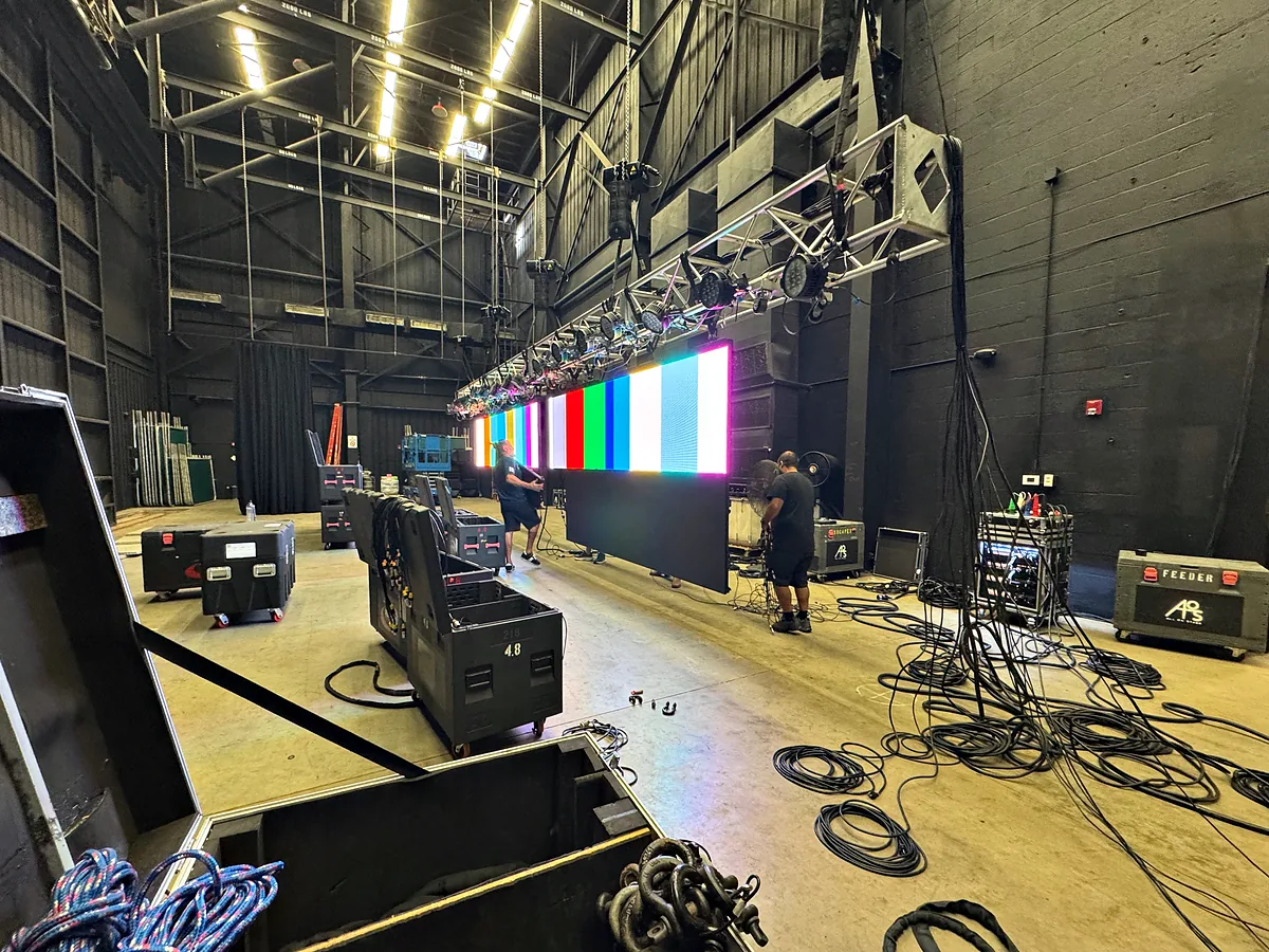 A video wall showing vertical color bars in a high-ceilinged hall, coils of cable and open road cases on the floor in front of it, loudspeakers on stands to the side and a truss of lamps overhead.