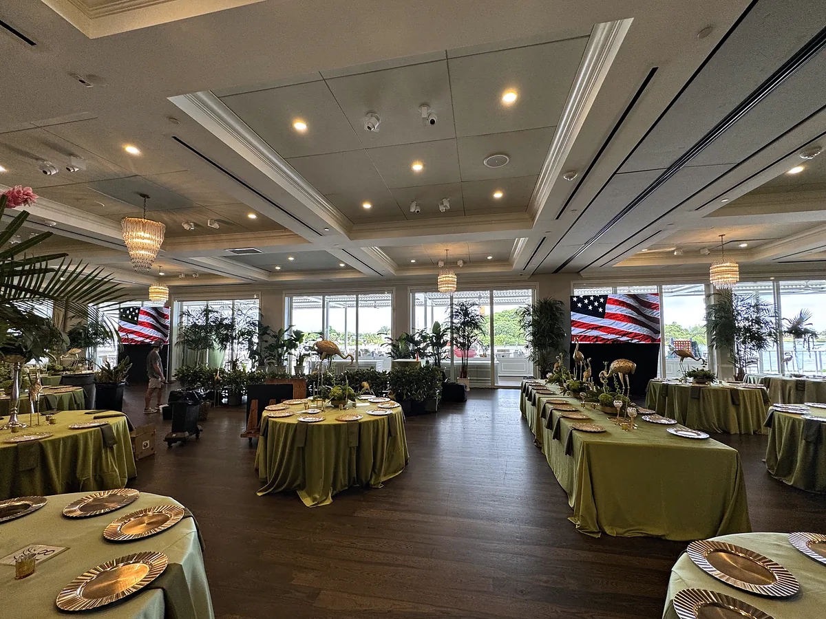 A room with windows along two sides set for dinner on round and long tables in green linen with gold chargers, planting between the tables, and a wide screen on the wall at each end carrying the same image.